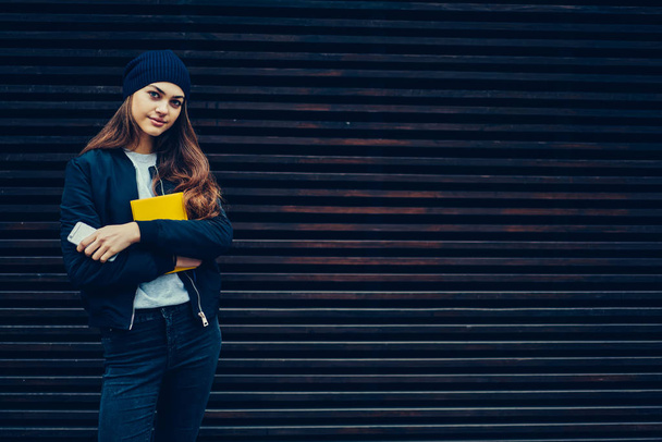 Portrait of teenager dressed in trendy clothing spending free time outdoors near to promotional background for your advertising message.Pretty student looking at camera while strolling at street - Φωτογραφία, εικόνα