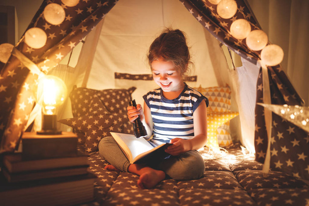 Счастливая девочка смеется и читает книжку в темноте в палатке у дома.
 - Фото, изображение