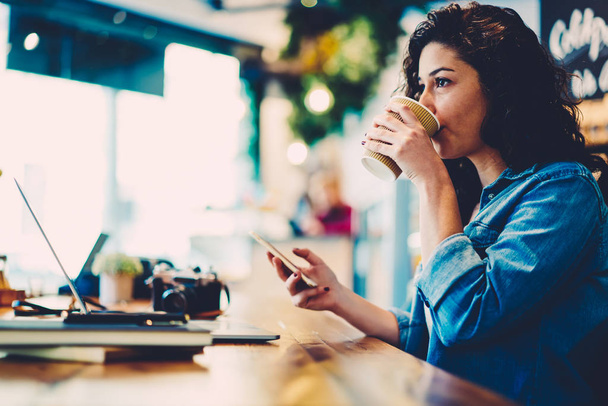 Attractive smart graphic designer drinking aroma coffee while distantly working on gadget in cafe.Young female person enjoying beverage while transferring money online via banking service on telephone - Fotó, kép