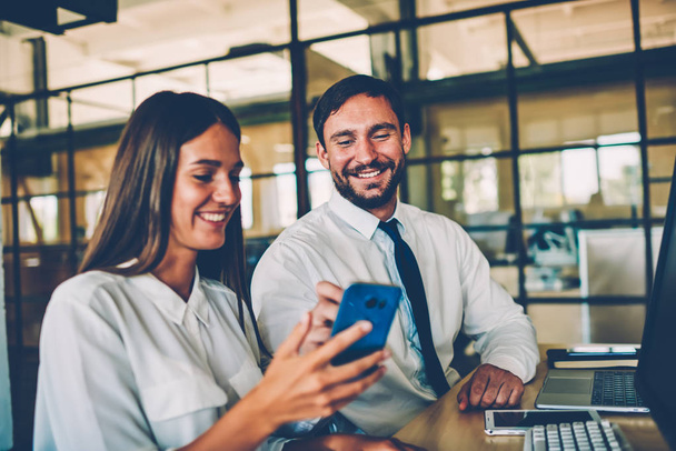 Cheerful female employee showing funny video on smartphone to her colleague during work break,coworkers having fun together in office reading news from networks and joking enjoying friendly talk - Photo, image