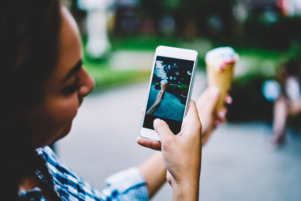Seleccione el enfoque en el teléfono digital moderno que sostiene a la mujer joven en la mano y hacer una foto de sabroso cono de helado para publicar post con comida en su propio blog caminando al aire libre en la calle de la ciudad moderna
 - Foto, imagen