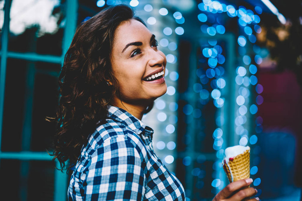 Side view of happy young woman with short curly hair and casual stylish shirt looking up and dreaming while holding ice cream in hand on colofrul promotion background enjoying recreation time - Fotó, kép