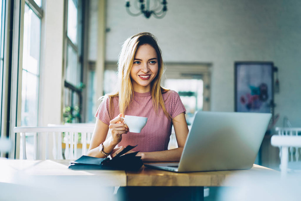 Прекрасная молодая владелица смотрит в сторону и улыбается, наслаждаясь кофе в кафе, работая с ноутбуком, подключенным к беспроводному интернету 4G.
 - Фото, изображение