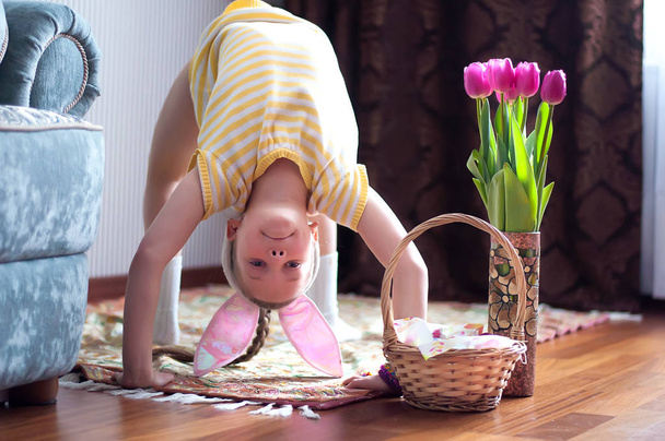 Симпатична маленька дитина носить вуха кролика на Великдень. Дівчина стоїть догори ногами біля кошика з розфарбованими яйцями
. - Фото, зображення