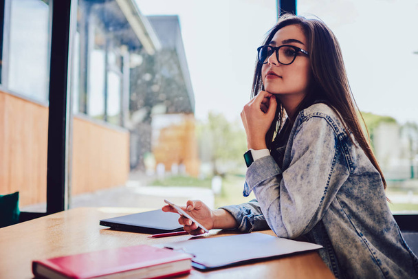 Contemplative student with cellular in hand looking away sitting at desk with textbooks.Copy space for your advertising text message.Thoughtful hipster girl in eyeglasses sending sms on telephone - Foto, afbeelding