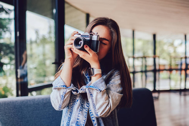 Professional female photographer making photos on vintage camera standing indoors in stylish coffee shop interior.Young woman focusing to taking pictures enjoying free time - Photo, image