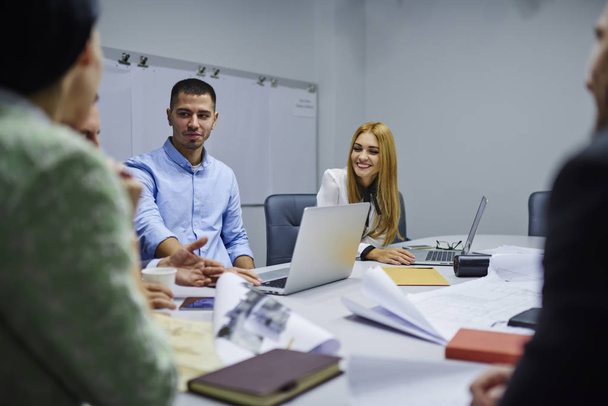 Команда улыбающихся профессионалов мужского и женского пола обсуждает новые идеи для развития финансовой компании и сотрудничает во время брифинга, сидя в офисном интерьере с современными цифровыми ноутбуками
 - Фото, изображение