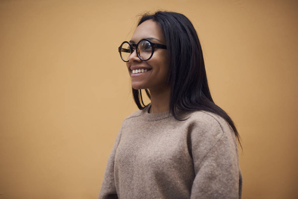 Beautiful smiling afro american female in spectacles standing on promotional background, attractive cheerful dark-skinned model in sweater enjoying free time dreaming about something on publicity area - Фото, изображение