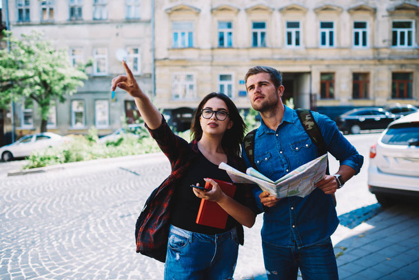 Young woman in eyeglasses and book in hands pointing direction to hansome male traveller with backpack.Female showing show place to tourist witch holding map in hands and searching destination - Foto, immagini