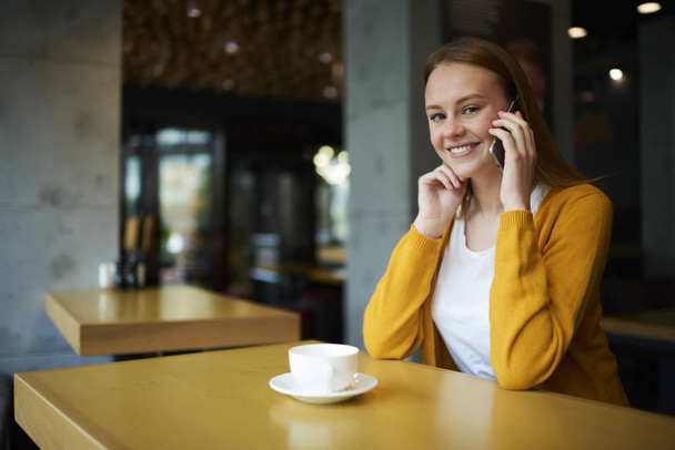 Portrait of happy businesswoman talking via modern smartphone using mobile benefits for roaming sitting in coffee shop and enjoying tasty cappuccino.Smiling female communicating on cellular - Φωτογραφία, εικόνα