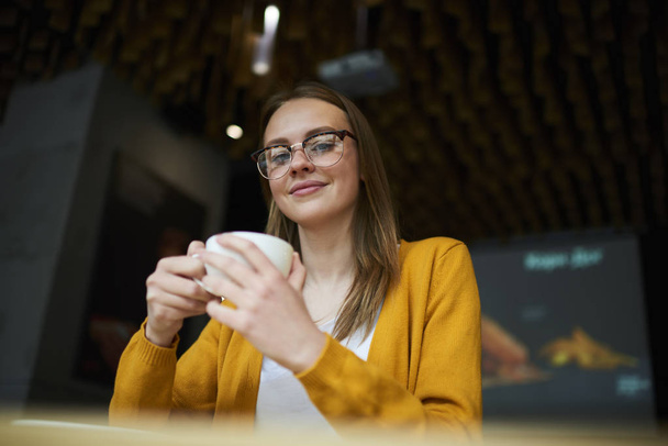 Portrait of good looking blonde hipster girl in stylish optical eyeglasses for better view holding cup of tasty coffee in hands and smiling at camera sitting in coworking space indoor - Фото, зображення