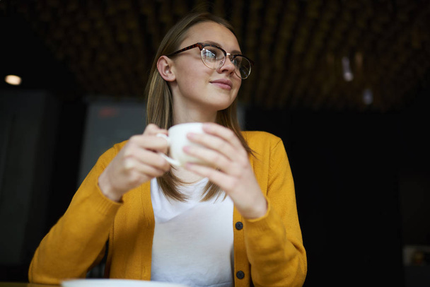 Beautiful female in trendy eyeglasses with good lenses looking away while holding cup of tasty coffee in hands.Attractive young woman with hot beverage spending free time in stylish cafeteria interior - Photo, Image