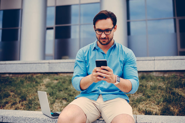 Young hipster guy in trendy eyewear chatting with friends via smartphone sending text messages, handsome student reading news from social networks while spending free time in university campus - Photo, Image