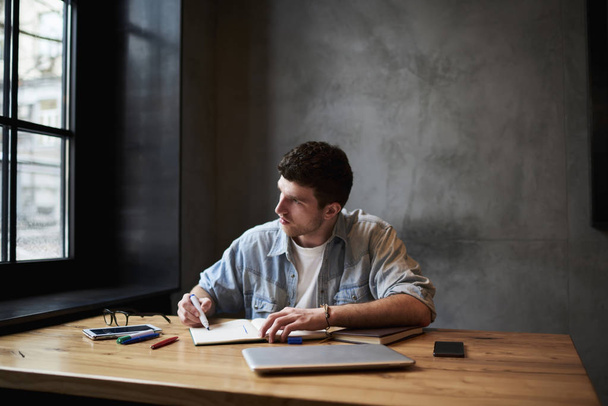 Молодой талантливый архитектор держит маркер в руке и рисует эскиз планирования здания в блокноте, глядя в окно, сидя за деревянным рабочим столом в интерьере студии во время рабочего процесса
 - Фото, изображение