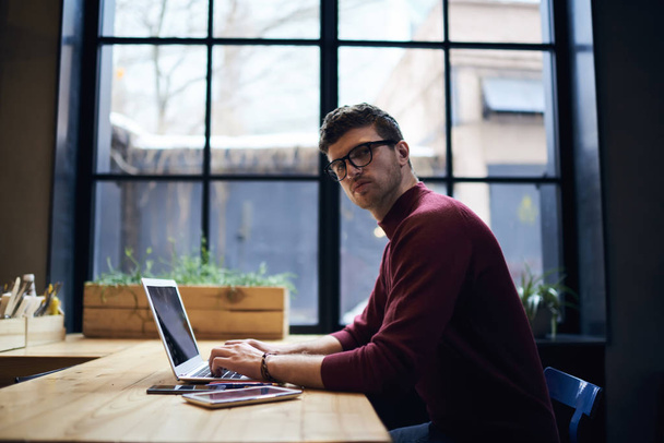 Portrait of skilled male student in optical eyeglasses working freelance at modern laptop.Young handsome entrepreneur looking at camera while keyboarding on computer device sitting indoors in office - Фото, зображення