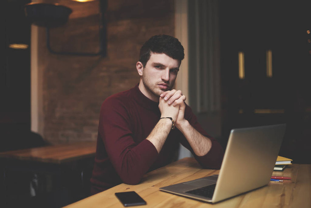 Half length portrait of confident entrepreneur looking at camera and pondering about good ideas for personal business project.Serious handsome website owner thinking while sitting at wooden table - Valokuva, kuva