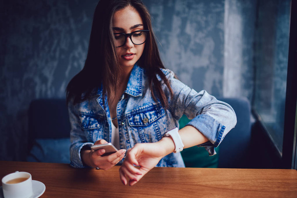 Busy female manager in stylish eyeglasses looking at watch while holding telephone in hand and hurry on meeting.Businesswoman keeping track of time sitting at table in coffee shop interior - Photo, Image