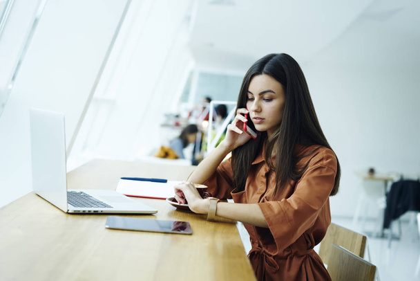 Charming concentrated female journalist checking time on watch planning working process sitting in coworking space, prosperous business woman arranging meeting while having phone conversation - Фото, зображення