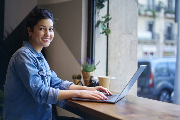 Portrait of cheerful young freelancer working at computer and using free high speed internet connection for searching information.Smiling student looking at camera while typing text on laptop device - Foto, Bild
