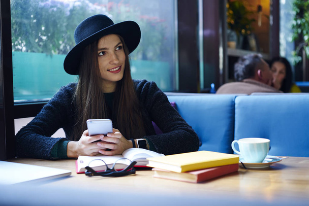 Attractive brunette hipster girl in trendy hat resting in cafe interior checking email on smartphone, smiling young woman blogging in social networks using modern mobile telephone on coffee break - Fotoğraf, Görsel