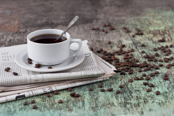 Чашка черного кофе на деревянном столе с разбросанными зернами и газетой. Черный кофе Горячий напиток Зерно Газета Деревянный фон Белый кубок Завтрак
 - Фото, изображение