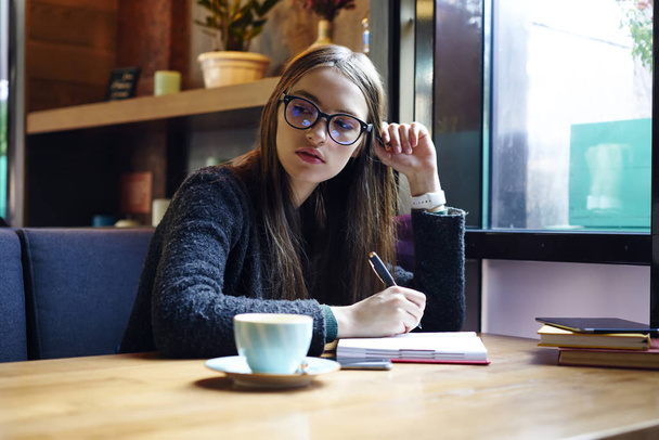 Pensive female student making report for homework thinking about accountings in cafe interior, clever graphic designer in eyewear pondering on creation sketch for project drawing in notebook - Фото, зображення