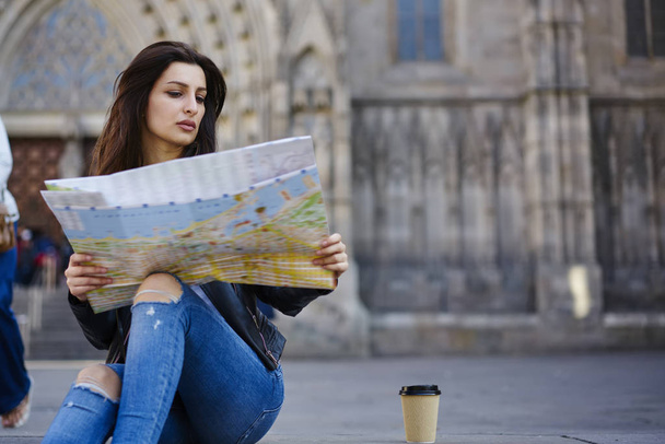 Concentrated brunette female traveler sitting on blurred urban setting background with map  searching route, tourist finding right direction on map for strolling in old europian city during vacations - Фото, изображение