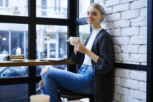 Portret van lachende blonde vrouwelijke student genieten van koffie tijdens pauze leren in moderne loft interieur café, aantrekkelijke lachende hipster meisje, drinken van thee doorbrengen vrijetijdsbesteding opstarten project planning  - Foto, afbeelding