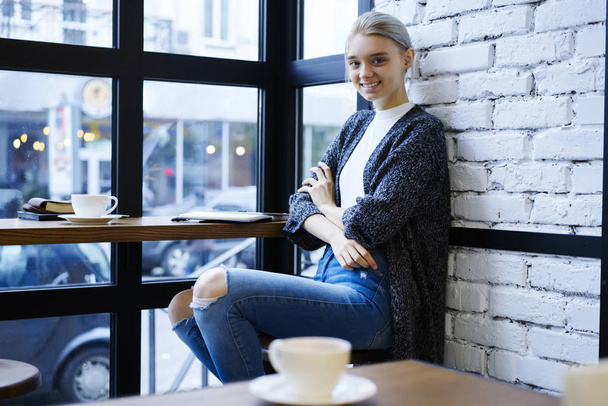 Attractive blonde hipster girl dressed in casual outfit sitting in coffee shop resting after working day,portrait of smiling student in enjoying free time sitting in modern loft interior cafe - Photo, Image