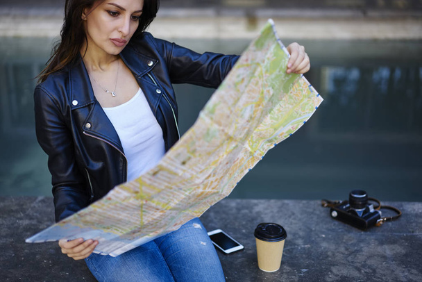 Konzentrierte junge Frau liest Landkarte und erschafft eine Art Stadtführung, während sie sich im Wochenendausflug erholt, attraktive Reisende suchen das richtige Reiseziel, um Orte in der Stadt zu zeigen, die auf städtischen Einstellungen sitzen  - Foto, Bild