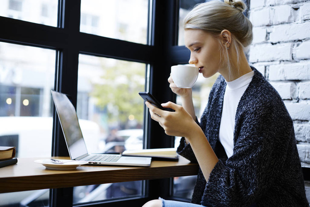 Atraktivní blondýna freelancer provádění nastavení smartphone při pití kávy na konec, mladý zkušený textař instalaci aplikace do mobilu pro synchronizaci souborů s přenosným počítačem během volného času - Fotografie, Obrázek