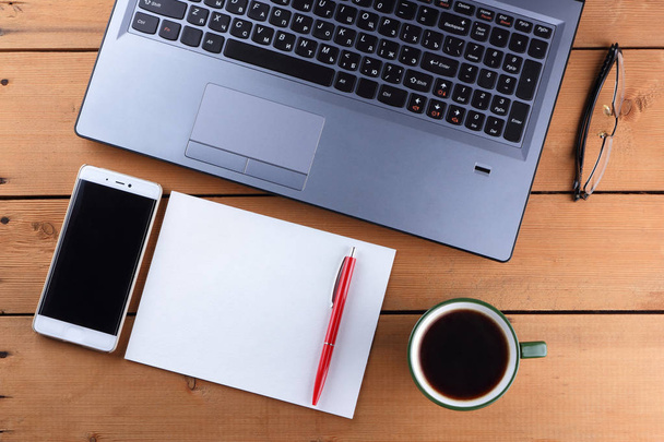 A laptop and a cup of coffee on a wooden background, a workstation on an old desk, a smartphone and a notebook, a pen and glasses in the workplace, a copy of space, a designer workstation in the minimalist style - Photo, Image