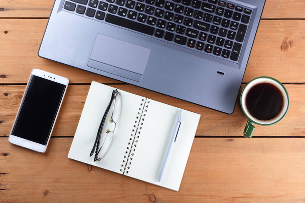 A laptop and a cup of coffee on a wooden background, a workstation on an old desk, a smartphone and a notebook, a pen and glasses in the workplace, a copy of space, a designer workstation in the minimalist style - Foto, Imagem