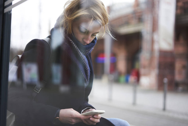 Pensive blonde traveler in coat and scarf using application on smartphone resting during city tour, concentrated hipster girl browsing information using 4G connection outdoors on advertising area - Fotoğraf, Görsel