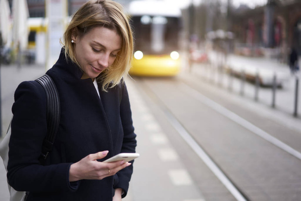 Positive blonde hipster girl reading news in social networks while waiting for tram on stop on blurred promotional background,attractive young woman using application monitoring public transport - Fotoğraf, Görsel