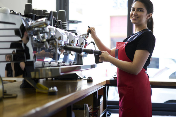 Portrait of cheerful attractive barista standing at modern automatic coffee maker with metal equipment and preparing tasty americano while looking at camera with smile and enjoying working process - Foto, imagen