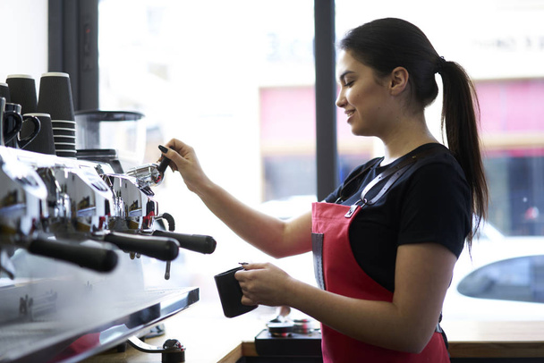 Positive young woman holding mug in hand and standing near modern espresso machine to preparing tasty coffee.Smiling waitress making hot beverage using automatic appliance in cafeteria interior - Foto, immagini