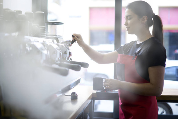 Professional  barista in uniform making aroma coffee using machine concentrated on working process.Beautiful brunette waitress preparing cappuccino doing order of cafeteria client standing at bar - Zdjęcie, obraz