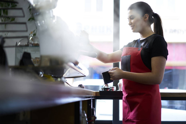 Attractive brunette female barista enjoying working process preparing coffee drinks using professional equipment in cafeteria.Skilled waitress in apron steaming milk on machine making cappuccino - Foto, immagini