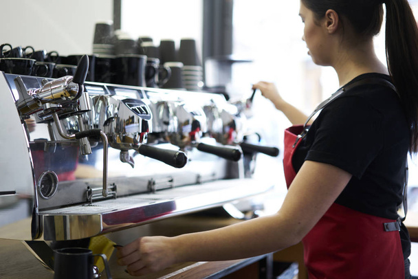 Cropped image of young woman working barista in coffee shop and preparing tasty espresso on modern coffee machine with metal professional equipment.Female waitress standing at automatic appliance - Foto, imagen