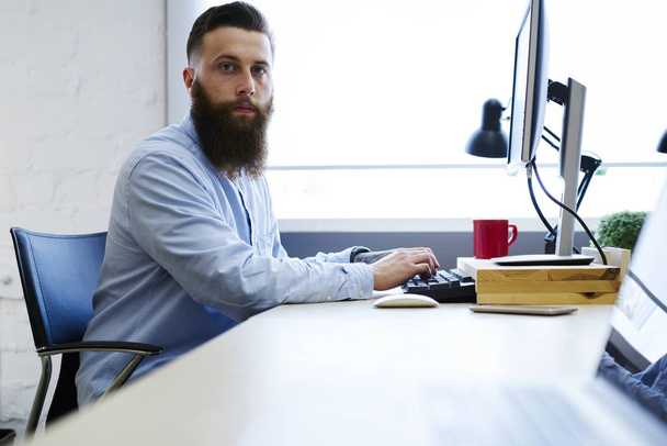 Half length portrait of  bearded employee looking at camera while browsing information in websites for work.Young hipster guy dressed in trendy clothing typing on keyboard sitting in office - Foto, immagini