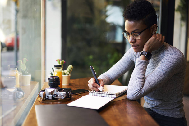 Attractive afro american female businesswoman concentrated on making planning of working schedule noting ideas while sitting in cafe doing remote job, talented young graphic designer drawing sketch  - Фото, зображення