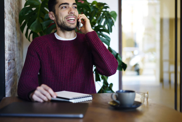 Cropped image of cheerful young man talking on modern cellular with friends sitting in coffee shop with cup of tasty beverage and notebook.Smiling guy having funny conversation on telephone - Фото, зображення