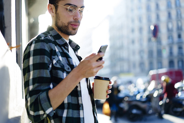 Σοβαρό όμορφος άνδρας στο θεάματα προσεκτικά παρακολουθώντας βίντεο στο τηλέφωνο συνδεδεμένο με 4g internet ενώ στέκεται σε ηλιοφάνεια αστικό σκηνικό. Ταξιδιώτη αναπαύεται με σύγχρονη συσκευή και καφέ - Φωτογραφία, εικόνα