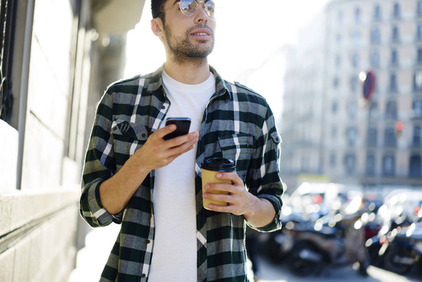 Όμορφος τουριστικός στο δροσερό μάτι γυαλιά αναζήτηση κατεύθυνση ενώ strolling στο δρόμο με το gadget και καφέ στα χέρια της πόλης. Γενειοφόρος άνδρας στοχαστικό χρησιμοποιώντας κινητό χάρτες σε αστικό περιβάλλον - Φωτογραφία, εικόνα