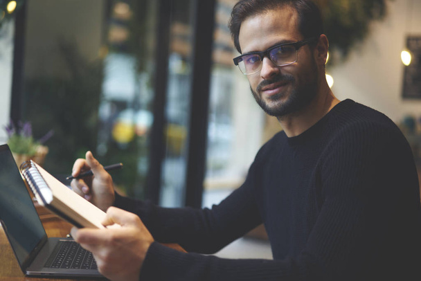 Portrait of bearded entrepreneur in stylish eyeglasses holding notepad and pen in hands and looking with smile at camera while working in coffee shop with computer device and using free internet - Photo, Image