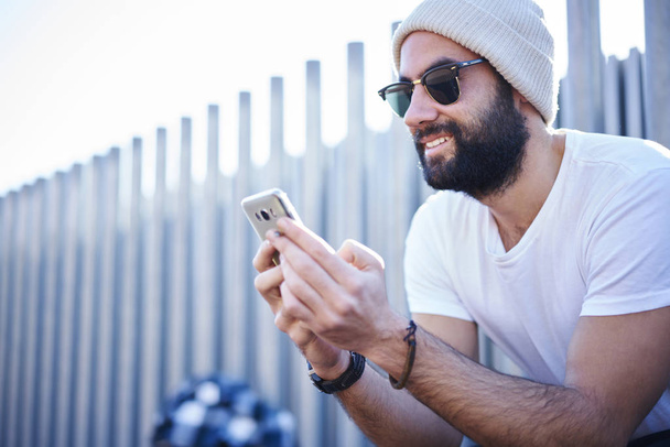 Cheerful young male hipster in trendy clothing and sunglasses watching funny video on modern smartphone enjoying free time outdoors sitting near copy space area for advertising on city street - Valokuva, kuva