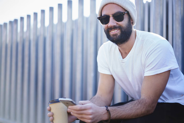 Portrait of cheerful bearded guy in trendy sunglasses sitting on urban setting promotional background looking at camera, trendy male hipster checking notification on smartphone texting messages - Valokuva, kuva