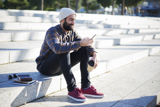 Sonriente chico hipster barbudo vestido con ropa casual leyendo noticias de la red social descansando al aire libre disfrutando del clima de primavera, turista masculino utilizando la aplicación en el móvil para navegar en la ciudad
  - Foto, Imagen