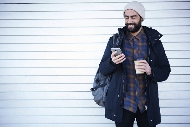 Positive young bearded guy dressed in casual outfit dialing number of taxi near copy space for advertising, male traveler standing with coffee to go using smartphone application for strolling - Fotó, kép